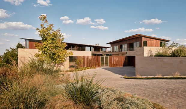 Wide exterior view of a contemporary multi-level residence with low, flat and gently pitched roof forms, warm timber cladding and vertical timber screening, pale masonry walls, expansive glazing, and a broad paved driveway. Drought-tolerant ornamental grasses and planting frame the foreground.