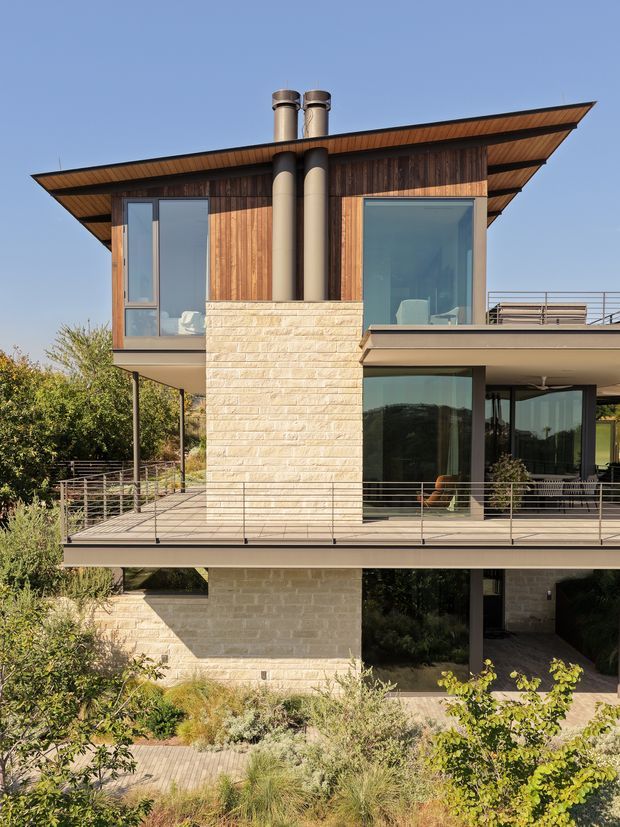 Exterior photograph of a contemporary multi-level residence built into a landscaped sloping site. The house features extensive floor-to-ceiling glazing, warm vertical timber cladding, pale horizontal stone masonry, broad cantilevered roof planes, large terraces with slim metal balustrades, and prominent cylindrical chimneys. Native-style planting surrounds the elevated structure.