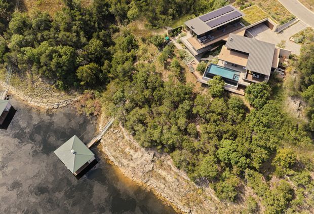 Aerial view of a contemporary multi-level residential property set among dense bushland on a rocky waterfront or lakeside edge. The complex has several connected low-pitched roof volumes, extensive outdoor terraces, and a rectangular swimming pool. A small roofed structure sits over the water below the property, with a road visible at the upper right.