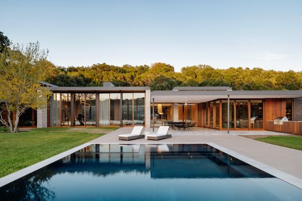 A single-storey contemporary residence with low, flat rooflines and extensive floor-to-ceiling glazing arranged around a large rectangular swimming pool. The home combines dark timber and neutral rendered or concrete-like exterior surfaces, with a broad paved terrace, landscaped lawn, mature trees, and a calm reflective pool in the foreground.