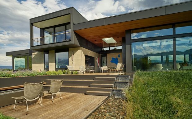 Modern multi-level home with strong indoor-outdoor connection, featuring dark metal cladding, pale masonry walls, extensive floor-to-ceiling glazing, a timber-lined soffit, and a broad timber deck stepping down toward landscaped planting. A covered outdoor dining and lounge area sits beside large glass openings, while the upper-level balcony has slim metal balustrades. Mountainous scenery is visible through the glazing and around the property.