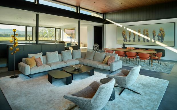A spacious, light-filled contemporary open-plan living and dining area with floor-to-ceiling glazing, a large sectional sofa, sculptural lounge chairs, a dark dining table with rust-coloured chairs, and a linear wall artwork. The room features a warm timber ceiling, dark vertical wall cladding, pale carpet, and expansive views through the glazing.