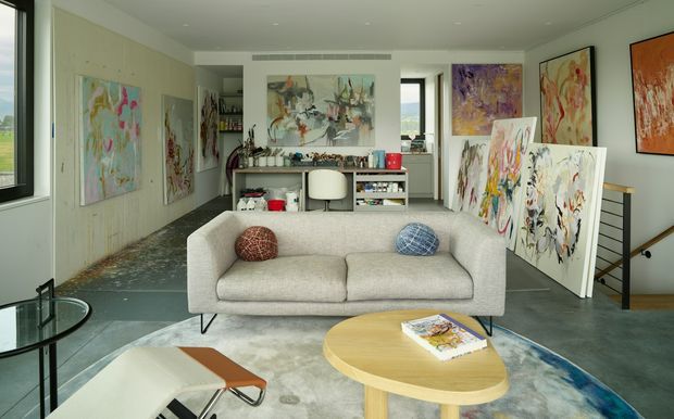 Bright contemporary artist's studio and living area with a neutral upholstered sofa, round timber coffee table, lounge chair, and extensive colourful abstract artwork displayed on the walls and leaning on easels. A compact work desk and shelving sit at the rear, with large windows, polished concrete flooring, and a stair opening visible to the right.