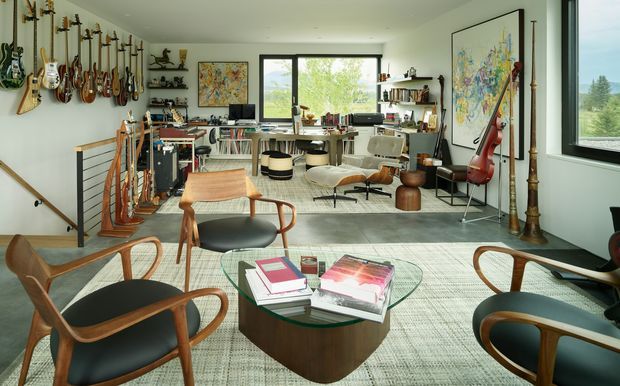 A bright, spacious music-focused living room or studio with guitars and string instruments displayed along the walls, a desk and computer workspace at the rear, lounge seating, a glass coffee table, framed artwork, shelving, and large windows overlooking greenery. The room combines mid-century furniture with eclectic artistic and musical character.