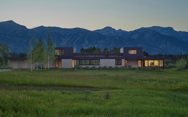 A wide exterior view of a long, low-slung contemporary residence set in an open grassy landscape, with dark rectangular volumes, extensive glazing, warm interior lighting, and a dramatic mountain range forming the backdrop at dusk.