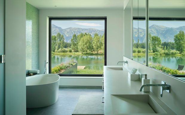 A contemporary minimalist bathroom with a freestanding white bathtub, double wall-mounted vanity basins, large frameless mirror panels, and expansive black-framed glazing overlooking a tranquil lake, trees, and mountains. The room has pale smooth surfaces, a light grey floor, clean geometric joinery, and abundant natural light.