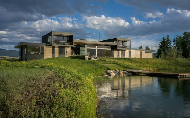 A modern, low-profile multi-wing house set in a broad grassy landscape beside a reflective pond. The building features rectilinear forms, large glazed openings, dark metal or charcoal cladding accents, pale masonry or concrete walls, and a mostly flat roofline. A small timber or dark-finished footbridge extends over the water in the foreground, with mountains and trees visible in the distance beneath a dramatic partly cloudy sky.