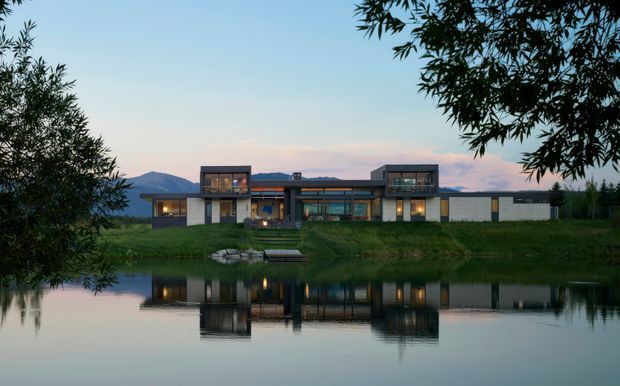 A contemporary low-rise residence viewed from across a calm pond at dusk. The long, horizontally composed house features dark framed glazing, projecting box-like upper volumes, pale wall panels, and warm interior lighting, with the building reflected in the water and framed by trees. Mountain silhouettes are visible in the background.