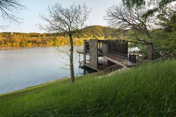A small contemporary timber-clad structure projects over the edge of a calm lake, accessed by a narrow ramp and elevated deck. The building is surrounded by mature trees and long grass, with wooded hills across the water. The scene appears to be a lakeside outdoor retreat or pavilion viewed from the grassy shore.