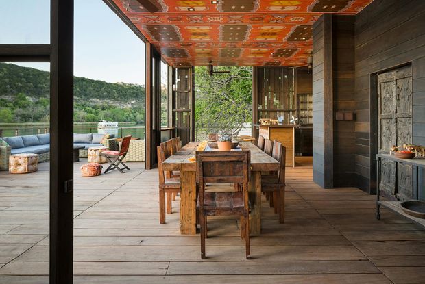 A rustic, open-plan dining area opening onto a waterfront terrace. The space features a substantial reclaimed timber dining table and chairs, dark horizontal timber wall cladding, broad timber flooring, extensive black-framed glazing, and a vividly patterned orange decorative ceiling. A fireplace or decorative built-in feature is visible along the right wall, while outdoor seating and a calm river or lake landscape are visible beyond the open glazing.