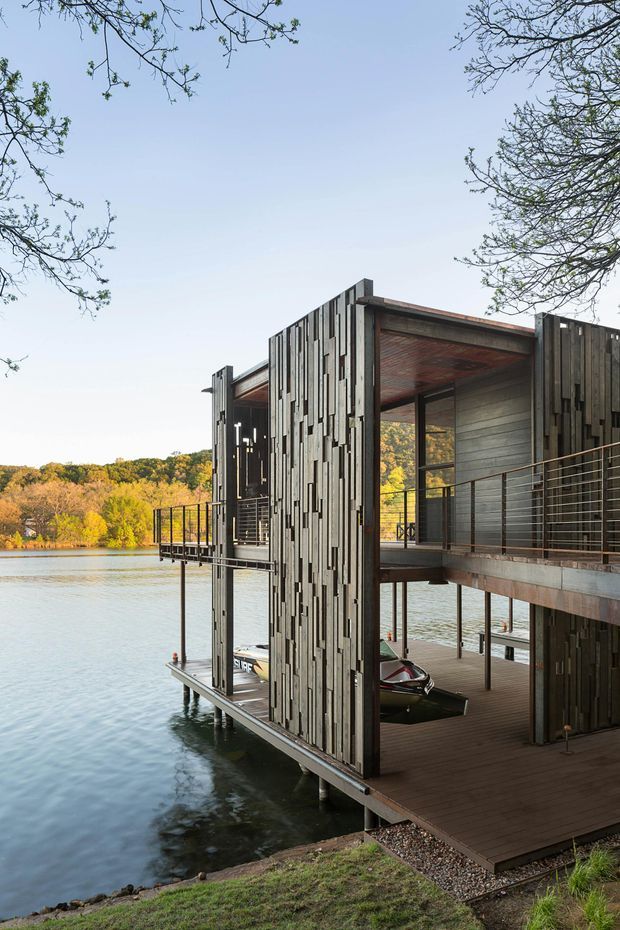 A contemporary multi-level waterfront structure extending over a calm lake, framed by trees and autumn foliage. The building features extensive dark timber cladding with varied vertical board patterns, open-sided decks, large glazed openings, exposed timber ceilings, and slim horizontal metal railings. A boat is moored beneath the elevated deck, reinforcing the lakeside setting.