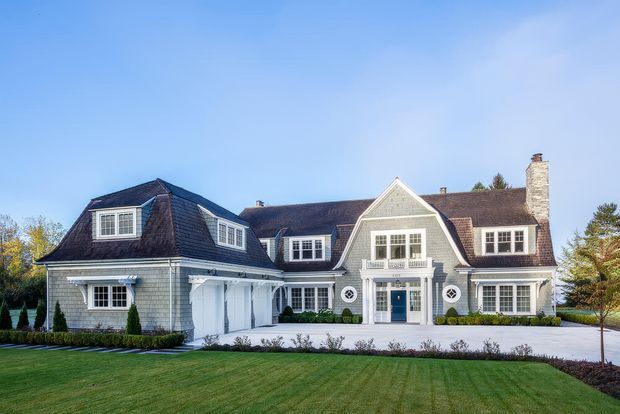 A large, symmetrical multi-wing residential house viewed from the front, set behind a manicured lawn and landscaped garden. The facade combines light grey shingle-style cladding, white trim, dormer windows, steep gabled roof sections, a central covered entry with balcony detail, attached garage doors, and a prominent stone chimney. The overall appearance is refined coastal-traditional and substantial in scale.