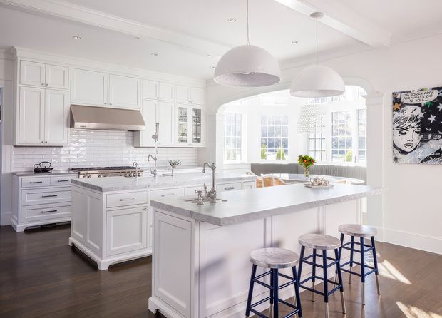 Bright, spacious white kitchen with classic raised-panel cabinetry, a large central island with a pale stone benchtop, tiled splashback, stainless steel rangehood and appliances, and three blue bar stools. Large multi-pane windows and an arched opening bring in abundant natural light. Dark timber flooring and oversized white pendant lights contrast with the predominantly white fit-out, while a bold artwork adds a small graphic accent.