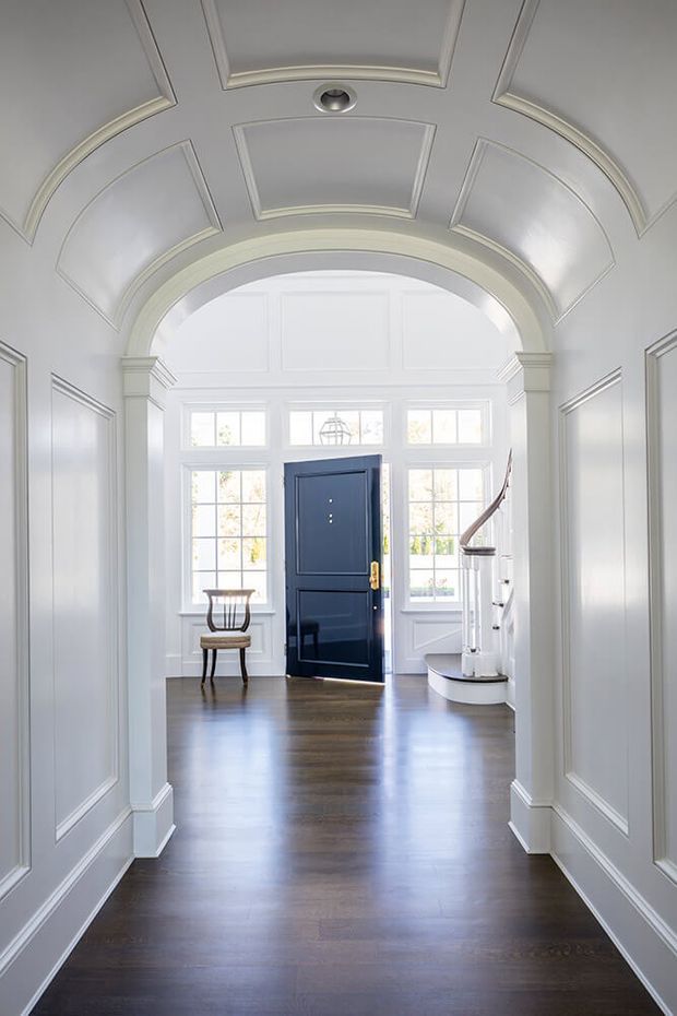 Bright, formal residential entry hall with extensive white-painted wall panelling, a decorative coffered and barrel-vaulted ceiling, arched opening with classical pilasters, dark timber flooring, and a navy blue front door surrounded by multi-pane windows. A curved staircase is visible at the far end, along with a small traditional chair.