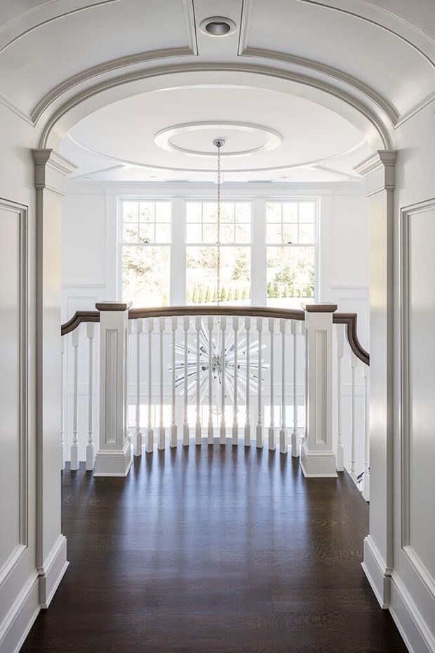 Bright, formal residential landing or upper-level hallway overlooking a curved staircase. The space features dark timber flooring, white painted wall panelling and elaborate moulded archways, columns, cornices and coffered ceiling details. A white turned-baluster balustrade with dark timber handrails curves around the stair opening, while large multi-pane windows flood the area with natural light. A central hanging light and recessed ceiling lights complete the refined traditional interior.