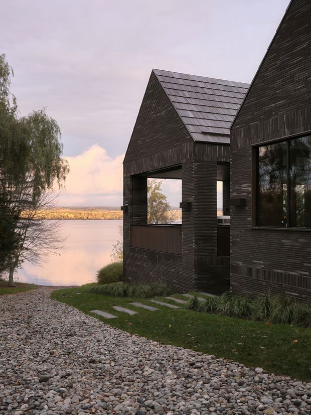 Exterior architectural photograph of a dark, linear-clad lakeside building with steep gabled roof forms, deep covered openings, and large dark-framed windows. A rocky path and landscaped lawn lead toward calm water, with trees and distant shoreline visible beyond. The subdued dusk light creates a quiet, contemplative atmosphere.