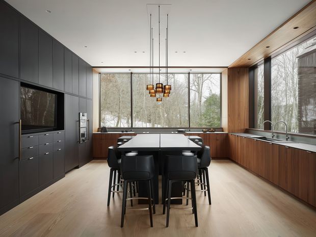 A spacious contemporary kitchen and dining area with a large central island and integrated seating for several people. The room features dark full-height cabinetry with built-in television and drawers, warm timber cabinetry and wall panelling, pale timber flooring, expansive floor-to-ceiling windows overlooking trees, and a cluster pendant light above the island.