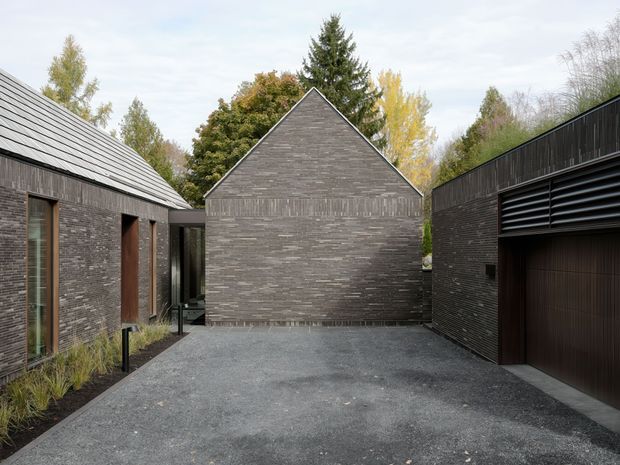 Exterior view of a contemporary dark-brick residential compound arranged around a gravel courtyard. The central gabled volume has a sharply pitched roof and textured horizontal brickwork, flanked by lower dark-brick buildings with metal roofing, narrow timber-toned openings, and a large louvred garage door. Sparse planting borders the courtyard, with mature trees visible beyond.