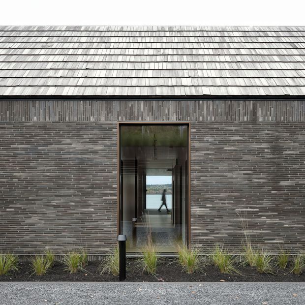 Front-facing exterior view of a low, highly textured building clad in narrow grey-brown masonry units, with a broad weathered metal shingle roof and a tall glazed entrance. The glazing provides a view through the building toward water and landscape, while sparse ornamental grasses sit in a dark planted strip along the facade.