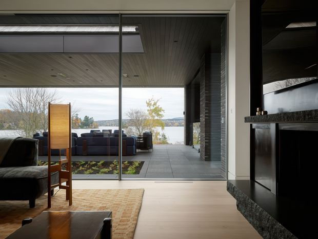 A refined contemporary living room opens through expansive floor-to-ceiling glazing onto a covered outdoor terrace overlooking a body of water. The room includes a dark upholstered sofa and armchair, a low timber coffee table, a patterned area rug, and a dramatic black fireplace surround with a textured stone ledge. The ceiling continues outward with linear timber boards, while the terrace uses large grey tiles and dark vertical screening.