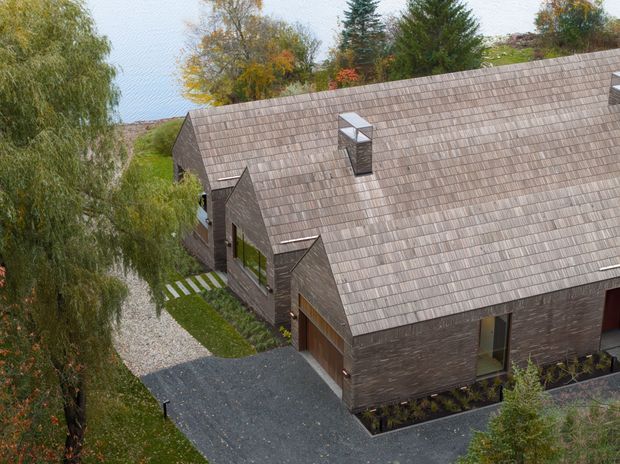 Elevated exterior view of a long, low-slung residential building with a steep pitched gable roof, dark weathered-looking cladding, and matching roof shingles. Multiple gabled projections, roof skylights, timber-framed openings, a gravel path, and a dark paved driveway are visible, surrounded by mature landscaped trees and autumn foliage.