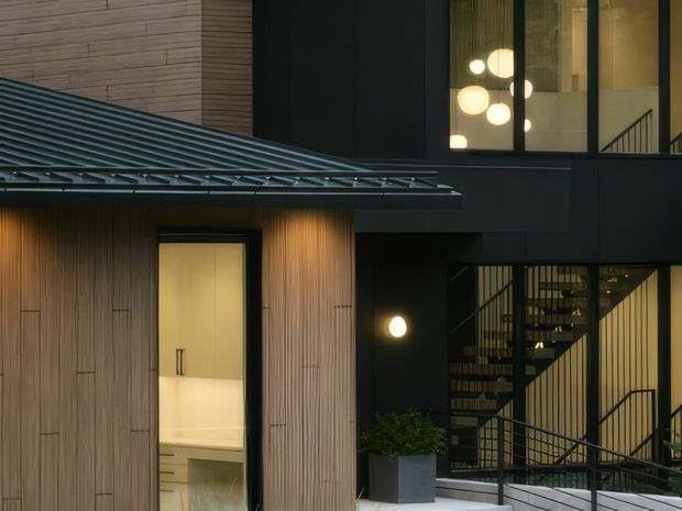 Exterior architectural photograph of a contemporary multi-level building at dusk. The facade combines dark smooth cladding with vertically grooved timber-like wall panels, a projecting corrugated metal roof, black-framed glazing, and an illuminated stairwell with black metal railings. Warm interior lighting and spherical pendant lights are visible through the windows, with a small planted container near the entry.