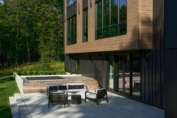 A contemporary multi-level home viewed from a landscaped outdoor terrace. The facade combines warm horizontal timber cladding, dark vertical cladding, and large black-framed windows. A paved patio with outdoor lounge seating sits beside raised timber-edged steps and a recessed lower-level glazed area, with mature woodland immediately beyond.