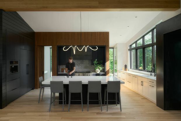 Contemporary open-plan kitchen and dining area with a large central island, four dark upholstered stools, extensive timber flooring, dark integrated cabinetry, and a warm timber feature wall around the cooking zone. Pale wood cabinetry runs beneath large black-framed windows on the right, bringing in abundant natural light. A sculptural linear pendant hangs above the island, with a living area or circulation space visible beyond the kitchen.