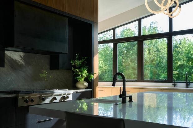 A sophisticated contemporary kitchen featuring dark timber and black cabinetry, a large stone island with an integrated sink and black gooseneck faucet, and a professional stainless-steel range. A full-height window wall brings in abundant natural light and views of greenery, while a sculptural ring pendant adds a decorative focal point.