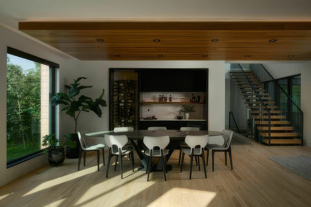 A contemporary open-plan dining area with a large dining table and mixed light chairs, positioned beneath a warm timber-lined ceiling with recessed downlights. The room features pale timber flooring, white walls, expansive black-framed glazing, indoor plants, and a built-in dark display and shelving unit. A partially open metal staircase with timber treads is visible to the right, creating a connection to the adjacent level.