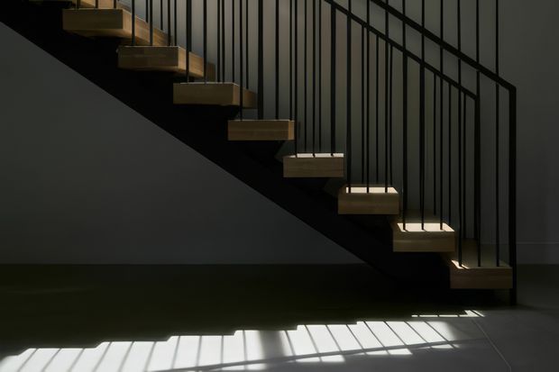 A dramatic, minimalist interior view of a floating staircase with light timber treads and slender black metal balustrade rods. The stair runs diagonally across a dark wall, with strong sunlight casting linear shadows across the tiled floor. The composition emphasises geometric forms, contrast, and the sculptural quality of the stair.