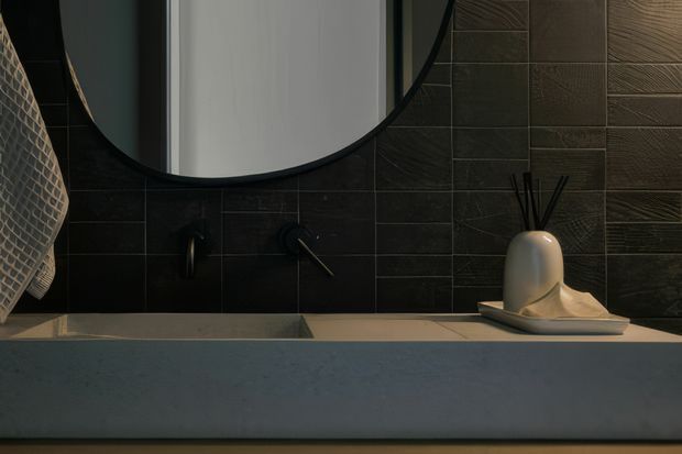 Close-up view of a dark, refined bathroom vanity with a large round black-framed mirror, wall-mounted black tapware, a pale stone or concrete benchtop, and dark textured rectangular wall tiles. A sculptural diffuser sits on the counter, with a woven hanging element partially visible at the left edge.