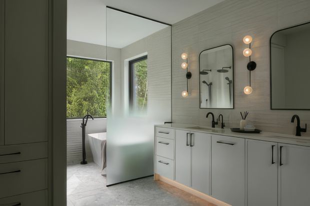 Contemporary bathroom with a long double-sink vanity, pale cabinetry, dark tapware, large mirrors, and globe wall lighting. A freestanding bathtub and additional fixtures are visible behind a frosted glass privacy partition. The room has pale horizontally textured wall surfaces, light stone-look flooring, and views to greenery through large windows.