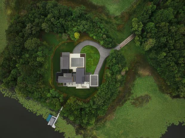 Aerial, top-down view of a contemporary house set within dense woodland and expansive landscaped grounds. The building has a clustered, rectilinear plan with several flat-roofed volumes, a curved driveway, and a small waterside structure or dock visible at the lower left.
