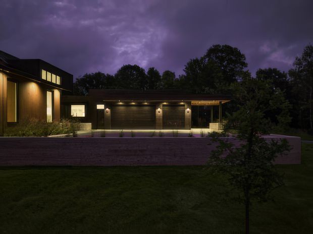 A dark, atmospheric dusk exterior view of a contemporary residential property. The composition shows a low, elongated garage or outbuilding with a warm illuminated entrance and garage doors, connected to a taller modern wing at left with large glowing windows. Timber-toned cladding, clean horizontal rooflines, recessed exterior lighting, a broad paved forecourt, and surrounding mature trees are visible.