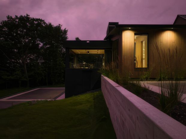Twilight exterior view of a modern single-storey residence, with dark vertical timber cladding, a flat roofline, warm recessed lighting, and large glazed openings. A low rendered masonry garden wall and landscaped grass and planting lead toward a covered outdoor living area, while mature trees frame the property.