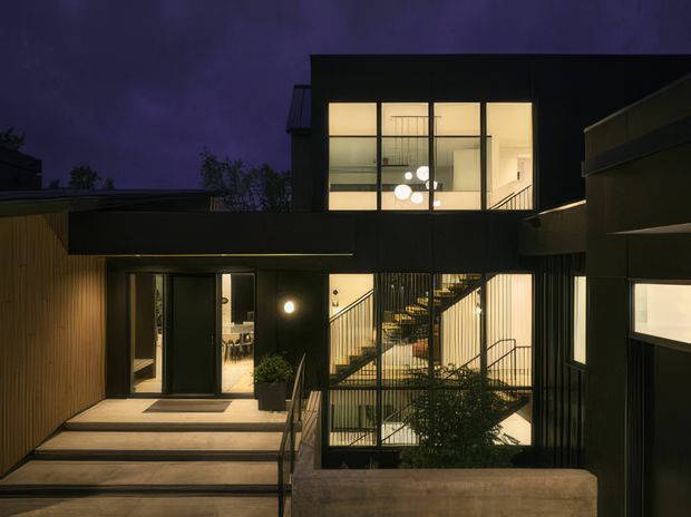 Night-time exterior view of a contemporary multi-level residence. The dark rectilinear facade combines vertical timber-like cladding, extensive illuminated glazing, recessed entry steps, and a prominent open stairwell enclosed by slender black metal balustrades. Warm interior lighting contrasts with the moody purple evening sky, while raised concrete planters and sparse landscaping frame the entrance.