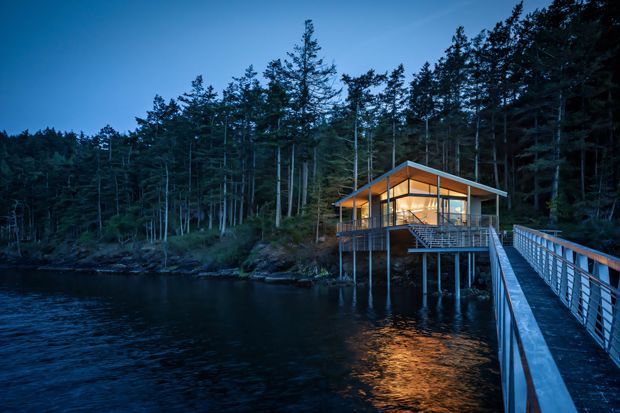 A contemporary pavilion-like house or retreat projects over a dark body of water at dusk, surrounded by dense forest. The structure has extensive floor-to-ceiling glazing, a low-pitched roof with prominent timber framing, and a raised deck supported on slender posts. A long pedestrian bridge with metal railings connects the building to the shore.
