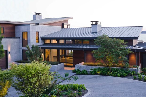 Exterior view of a sophisticated contemporary multi-level residence with low-pitched metal roofs, concrete walls, dark timber cladding, extensive glazing, and carefully landscaped garden beds. The entrance and several warm-lit interior spaces are visible through the glazing, while the main subject is the building facade and roof composition.