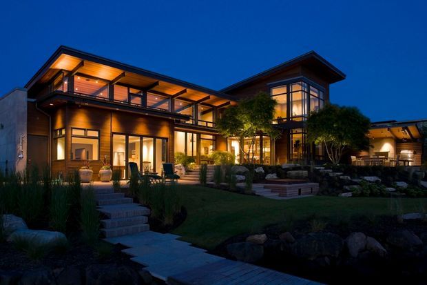 Twilight exterior view of a large multi-level contemporary residence with extensive warm-lit glazing, dark timber or wood-toned cladding, projecting roof planes, balconies, and landscaped outdoor terraces connected by stone steps and paths.