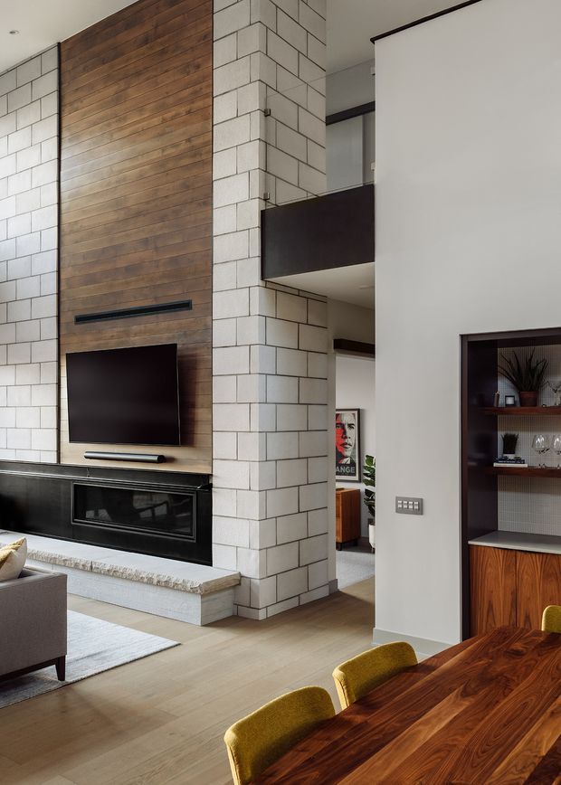 A contemporary open-plan living and dining area with a dramatic double-height central fireplace wall. White blockwork masonry forms the fireplace surround and structural column, contrasted with a warm vertical timber feature wall, black built-in fireplace and television. A timber dining table with mustard upholstered chairs occupies the foreground, while a dark built-in display cabinet and glimpses of adjacent rooms appear to the right and rear.