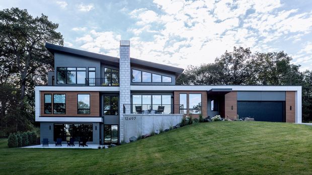 A contemporary multi-level residence set into a gently sloping landscaped site. The house combines dark metal roof planes, extensive black-framed glazing, warm timber-clad wall sections, pale masonry, and a prominent white brick chimney. A broad integrated garage sits on the right, while layered terraces and large windows define the facade. Mature trees and a manicured lawn surround the building.