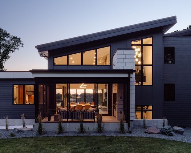 Twilight exterior view of a contemporary multi-level residence with dark horizontal cladding, broad glazed openings, a projecting upper volume, and a covered outdoor terrace. Warm interior lighting reveals living and dining areas through the glazing. The landscaped foreground uses gravel, sparse ornamental planting, and low concrete retaining elements.