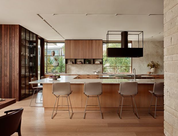A warm, open-plan contemporary kitchen viewed from the living area, featuring a long timber island with seating for five, integrated cabinetry, a stone-look splashback and benchtop, expansive windows, dark timber-framed glazing, and a sculptural suspended rangehood. A dining area and glazed display joinery are visible to the left.