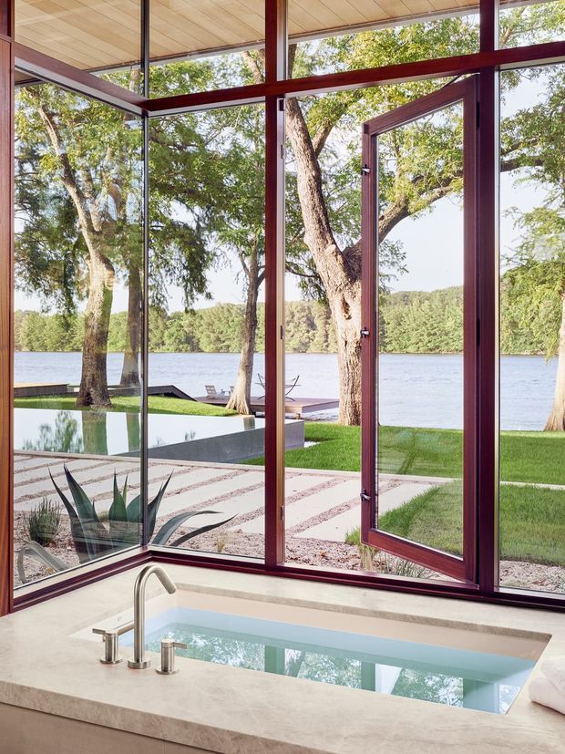A bright modern indoor spa or pool room with a small rectangular plunge pool set into a pale stone surround. Floor-to-ceiling glazing and an open glass door frame views of a landscaped lawn, trees, dock, and lake beyond. Dark reddish timber or metal window and door frames contrast with the light ceiling and stone surfaces.