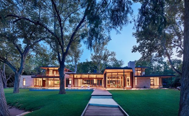 A wide exterior photograph of a low-rise modern residence set within a spacious, maturely landscaped garden. The home has a strongly horizontal composition, extensive floor-to-ceiling glazing, warm illuminated interiors, pale masonry walls, dark roof planes and a straight timber-and-paver pathway leading across the lawn to the entrance. Large established trees frame the house and create a serene, park-like setting.