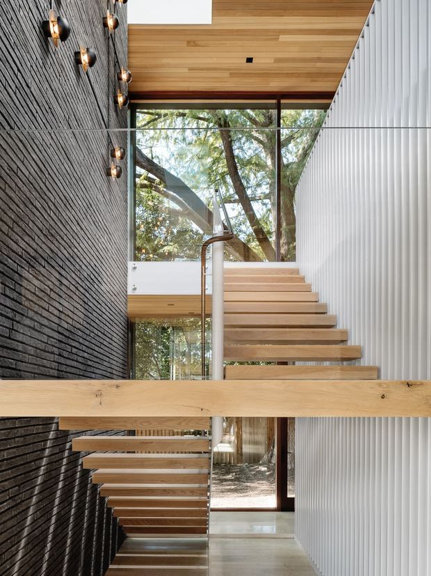 A contemporary interior stairwell with a floating timber staircase and glass balustrade. The space features a dark textured brick wall with sculptural wall lights on the left, a white vertically ribbed wall on the right, extensive glazing overlooking trees, warm timber ceiling boards, and a strong horizontal timber beam crossing the foreground. The open-riser stair visually connects multiple levels and creates a light-filled architectural feature.