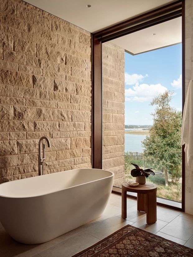 A warm, refined bathroom featuring a freestanding white soaking bathtub beside a tall dark-framed glazed opening. The walls are clad in rough-textured beige stone masonry, with light tiled flooring and a minimalist dark timber side table holding a plant and decorative objects. A sculptural gooseneck tap is mounted beside the tub. Through the glazing, a calm waterside landscape and blue sky are visible. The room has a contemporary natural-material aesthetic with strong indoor-outdoor connection.
