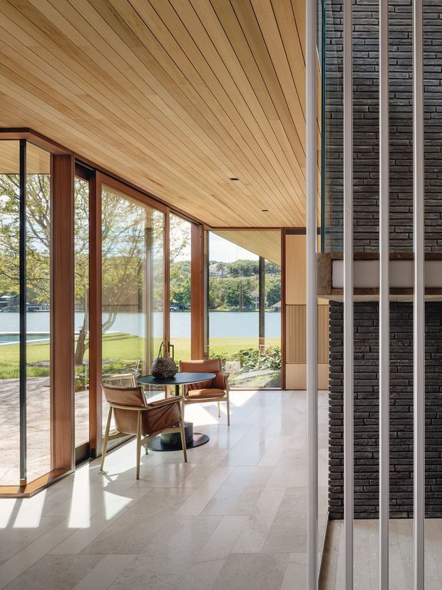 A refined, light-filled interior sitting area beside a waterfront, with full-height timber-framed glazing, a warm timber-lined ceiling, pale stone or tile flooring, and a dark textured brick feature wall with integrated shelving. Two leather lounge chairs and a small round table create an intimate seating nook. The space has strong indoor-outdoor connection and views across the water.
