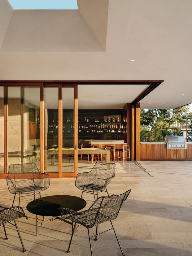 A spacious indoor-outdoor entertaining area with a covered terrace, opening through full-height timber-framed glazing to a dark built-in bar and dining counter. The terrace has pale stone paving, black wire-frame outdoor dining furniture, timber cabinetry, and a built-in stainless-steel barbecue. A large skylight and recessed ceiling lights brighten the space, with mature trees visible beyond.